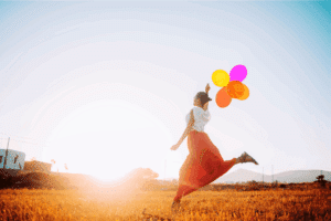 Frau springt auf einer Wiese mit bunten Luftballons – Symbol für Leichtigkeit, Freude und Glück.